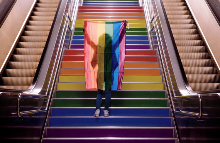 Escada com as cores do arco-íris, e pessoa em frente dela, levantando a bandeira LGBT