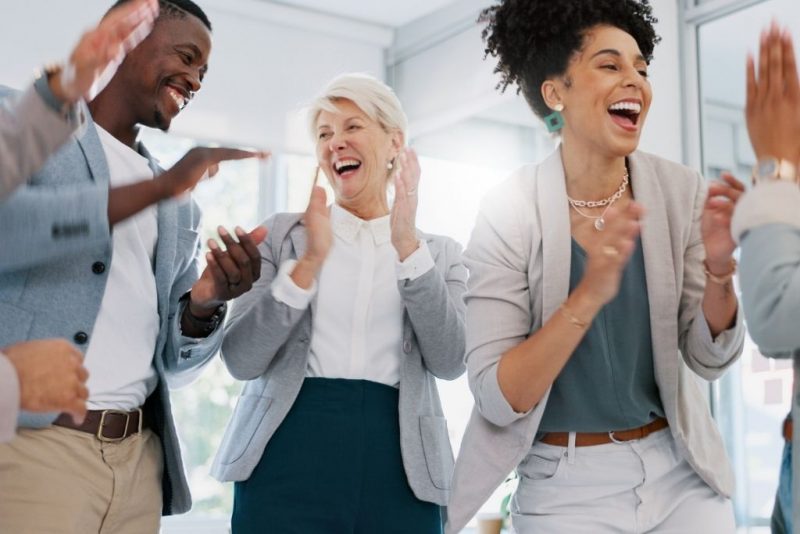 Profissionais de diferentes idades e etnias se divertindo e celebrando juntos em um ambiente de trabalho, simbolizando a importância de criar um clima organizacional positivo com a ajuda de benefícios flexíveis, que contribuem para a satisfação e motivação dos colaboradores.