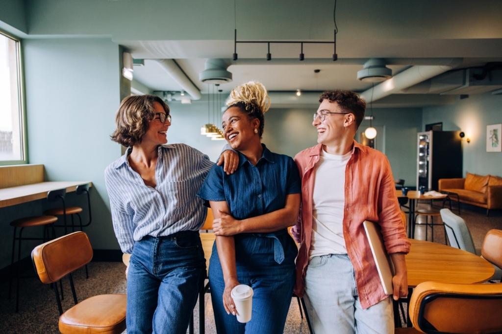 Três jovens profissionais sorrindo e interagindo em um ambiente de trabalho moderno, destacando um cenário colaborativo. A imagem reflete o impacto positivo que benefícios flexíveis podem ter na motivação e engajamento dos colaboradores, criando um ambiente mais dinâmico e saudável.