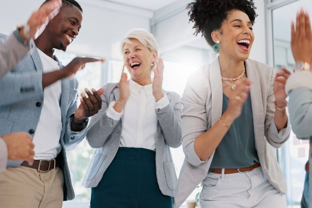 Profissionais de diferentes idades e etnias se divertindo e celebrando juntos em um ambiente de trabalho, simbolizando a importância de criar um clima organizacional positivo com a ajuda de benefícios flexíveis, que contribuem para a satisfação e motivação dos colaboradores.