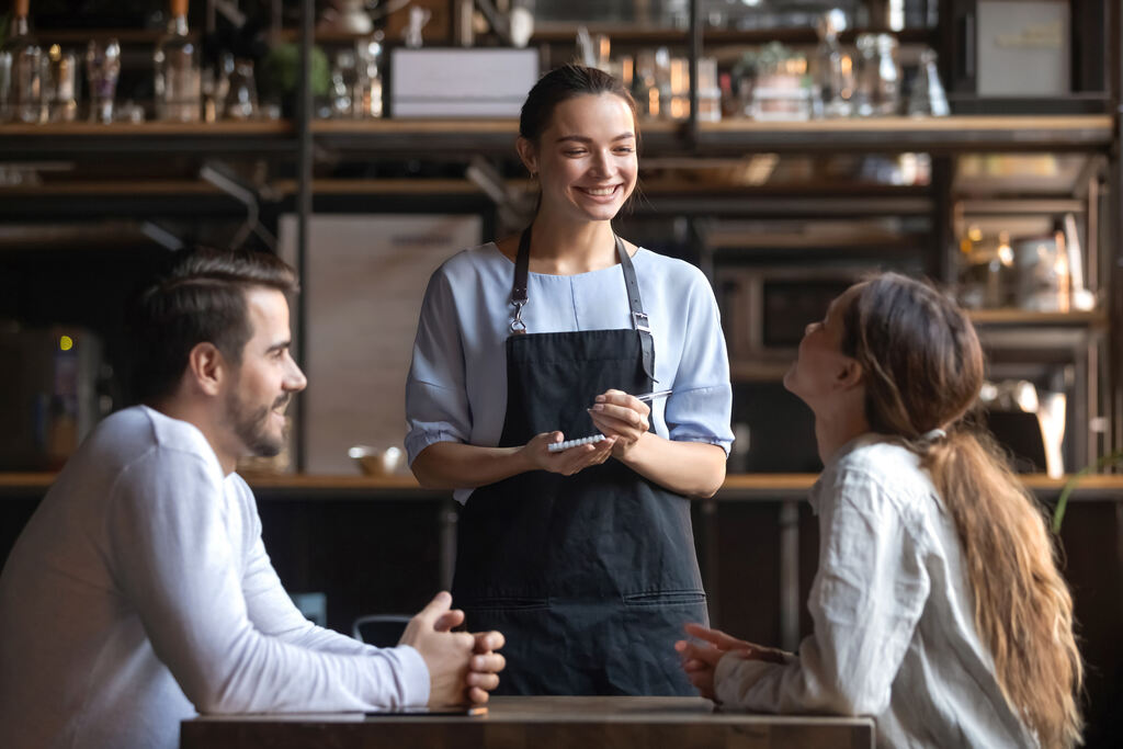 Em um ambiente interno de um restaurante ou cafeteria com prateleiras de garrafas ao fundo, uma garçonete sorridente de avental preto e blusa azul anota o pedido em um bloco de notas. Ela está posicionada entre um homem e uma mulher sentados à mesa, que a observam atentamente. A imagem transmite um clima de hospitalidade e serviço de alimentação pronta, contexto ideal para explicar a diferença entre vale alimentação e vale refeição quanto ao local de uso desses cartões.