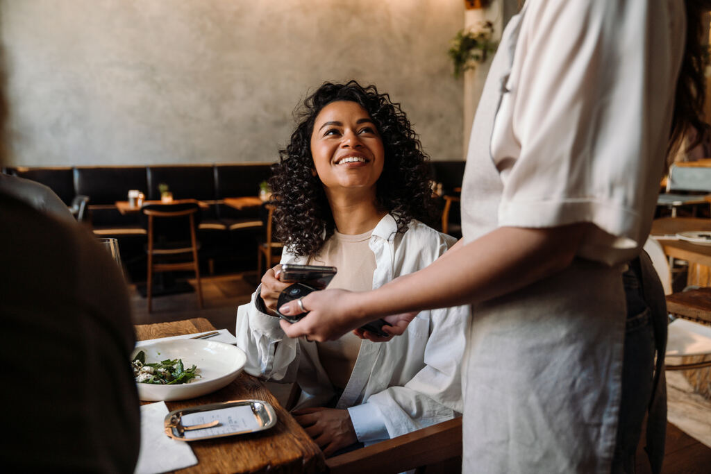 Uma mulher negra com cabelos cacheados e sorriso radiante está sentada à mesa de um restaurante. Ela olha para cima, em direção à atendente que está de pé, enquanto aproxima seu smartphone da maquininha de cartões para realizar o pagamento por aproximação. Sobre a mesa de madeira, há um prato com salada e uma pequena bandeja metálica com a conta. O cenário ilustra um momento de uso prático dos benefícios corporativos, ressaltando a diferença entre vale alimentação e vale refeição no cotidiano do trabalhador que opta por comer fora.
