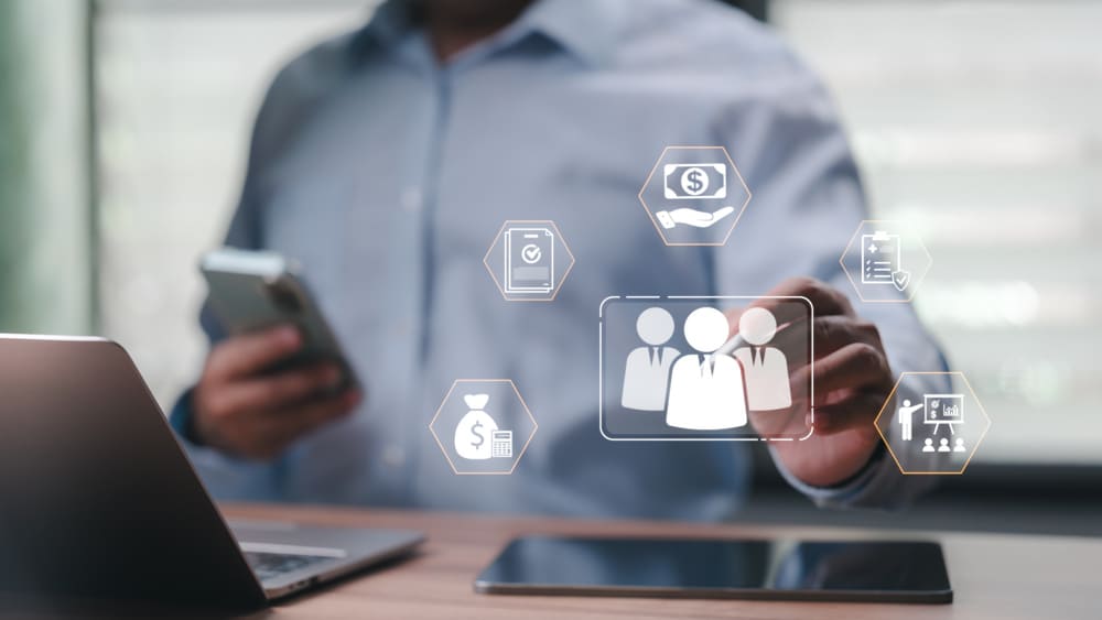 Homem utilizando notebook e smartphone enquanto interage com ícones digitais de gestão de pessoas, remuneração e recursos humanos, ilustrando a gestão de benefícios para pequenas empresas por meio de tecnologia e processos integrados.