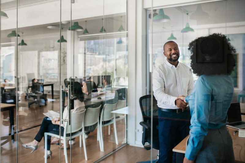 Homem e mulher apertando as mãos em escritório de empresa, com outros candidatos à vagas esperando em cadeiras.