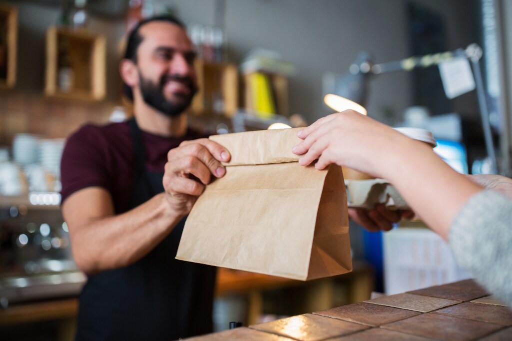 Funcionário entregando um pacote de comida para um cliente, ilustrando o uso do vale-refeição para empresas em estabelecimentos de alimentação, destacando a aceitação e conveniência do benefício.