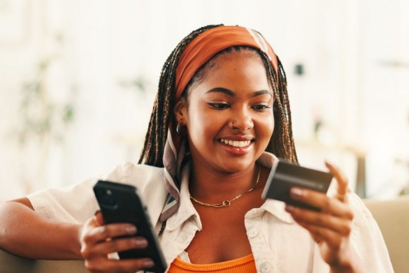 Mulher negra sorrindo enquanto segura seu celular em uma mão e um cartão de benefícios personalizados na outra mão, olhando para o cartão. A cena retrata um momento de satisfação ao gerenciar seus benefícios, demonstrando a facilidade de uso de um cartão de benefícios flexível.
