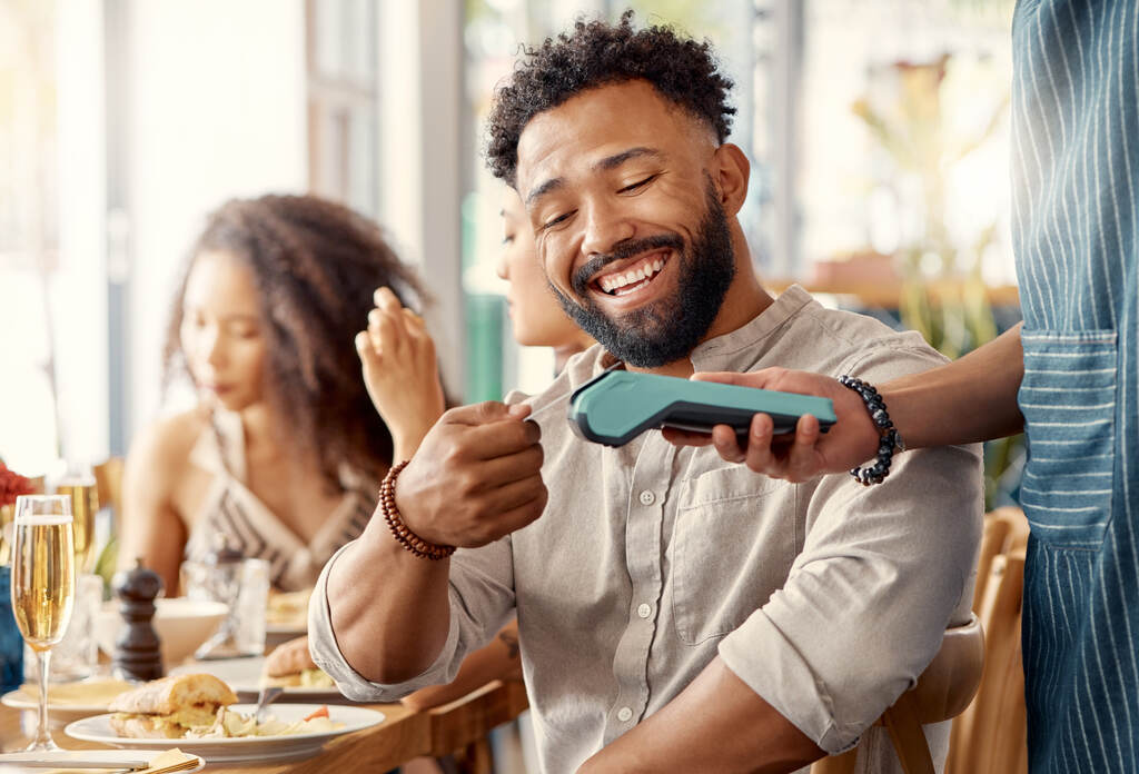 Homem sorrindo em um restaurante enquanto faz um pagamento com um dispositivo de pagamento sem contato. Ele está usando um cartão de benefícios personalizados, possivelmente para pagar a refeição, em um ambiente social e descontraído.