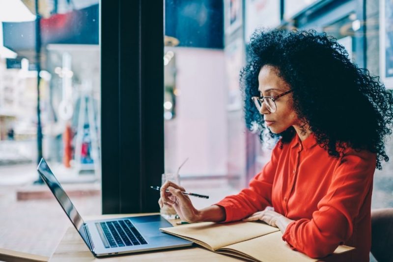 Mulher negra com cabelo cacheado e óculos, trabalhando sozinha em um ambiente de café, usando um laptop e anotando em um caderno, refletindo sobre os desafios do RH moderno, como a adaptação a novos modelos de trabalho.
