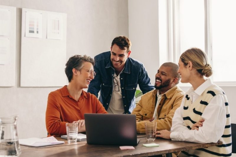 Quatro colaboradores estão sentados ao redor de uma mesa, interagindo e sorrindo durante uma reunião de trabalho. A imagem demonstra como uma política de benefícios eficaz pode aumentar a colaboração e a motivação dentro da empresa, favorecendo a criação de um ambiente saudável e produtivo.