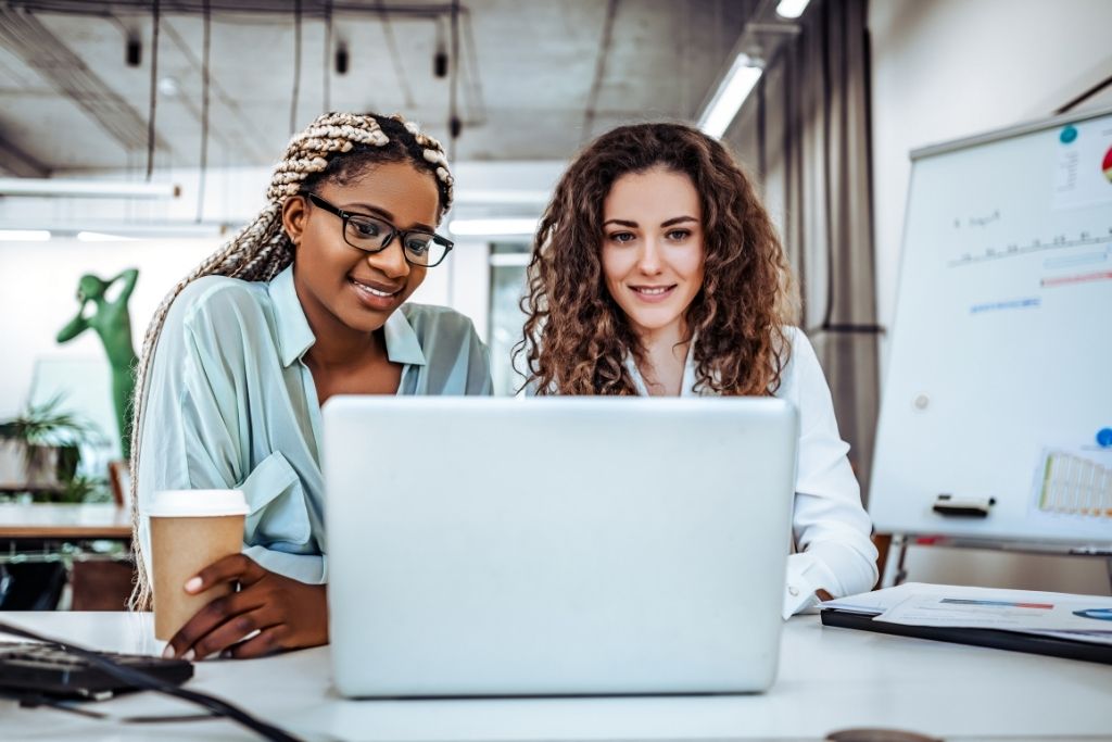 Imagem de duas profissionais de RH sorrindo enquanto trabalham juntas em um escritório moderno. Uma delas segura um copo de café e ambas estão usando um computador portátil. A imagem ilustra um ambiente de trabalho colaborativo e conectado, onde uma boa política de benefícios pode promover o engajamento e a satisfação dos colaboradores.