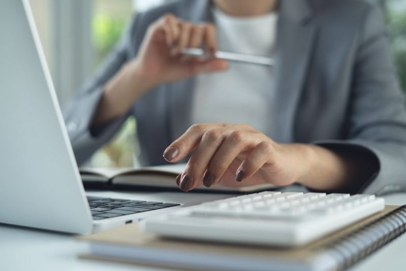 Close em uma mão de uma profissional, com unhas esmaltadas de marrom, interagindo com o teclado de um laptop e uma calculadora sobre um caderno. A imagem ilustra o desafio do RH em relação à redução de custos na gestão de benefícios, sem deixar de gerar valor aos colaborares e à empresa.