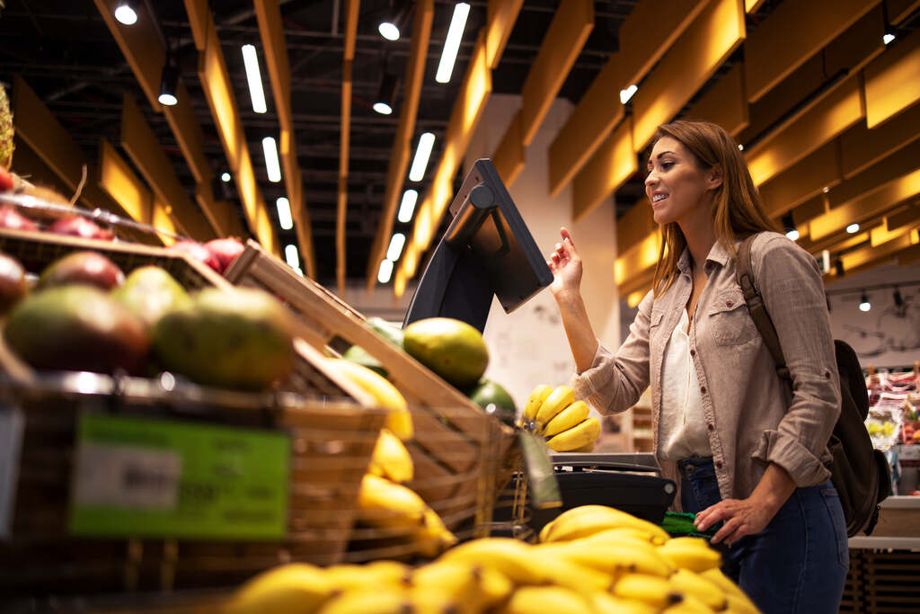 Mulher sorridente utilizando vale alimentação para pagar compras de frutas em mercado.