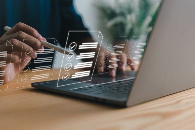 Imagem de uma pessoa interagindo com um laptop enquanto revisa documentos e listas digitais, representando a automação de processos no RH. A imagem simboliza a eficiência que a automação de processos pode trazer para as operações de Recursos Humanos ao facilitar a organização de tarefas e a gestão de dados de maneira digital e eficiente.