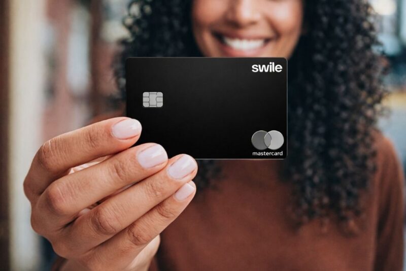 Uma pessoa sorridente segura em primeiro plano um cartão flexível da Swile. O cartão é totalmente preto com acabamento fosco, apresentando um chip eletrônico no lado esquerdo, o logotipo da Swile no canto superior direito e a bandeira Mastercard no canto inferior direito. O foco da imagem está no cartão, enquanto o rosto da mulher ao fundo aparece levemente desfocado.