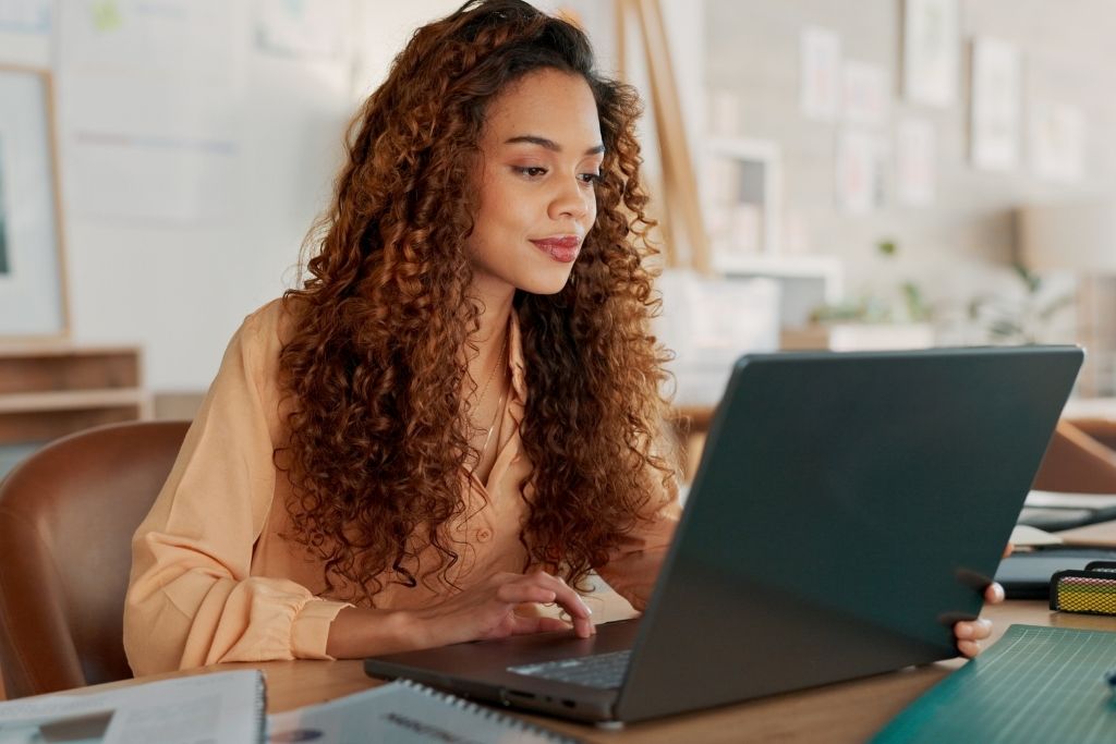 Uma mulher com cabelos cacheados está sentada à mesa em um ambiente de escritório bem iluminado, concentrada enquanto utiliza um notebook. A cena representa o processo de gestão ou contratação do cartão flexível por parte de uma empresa, destacando um ambiente de trabalho moderno, organizado e produtivo.
