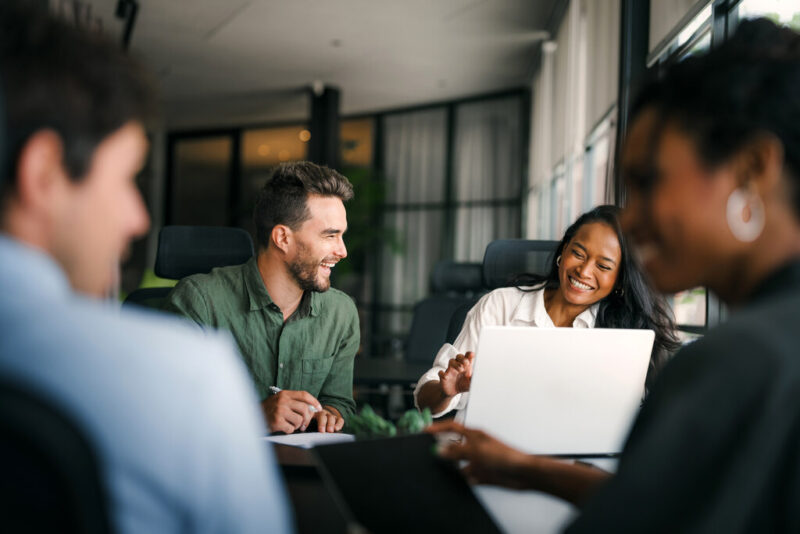 A imagem apresenta quatro colegas de trabalho em uma reunião descontraída dentro de um escritório moderno, exemplificando um clima organizacional positivo e colaborativo. No centro, um homem branco de camisa verde sorri abertamente enquanto segura uma caneta, olhando para uma colega negra que também sorri enquanto utiliza um notebook branco. Em primeiro plano, as silhuetas de outros dois colegas embaçados emolduram a cena, criando uma sensação de profundidade e inclusão. O ambiente é iluminado por grandes janelas ao fundo, reforçando a ideia de transparência e bem-estar no trabalho.