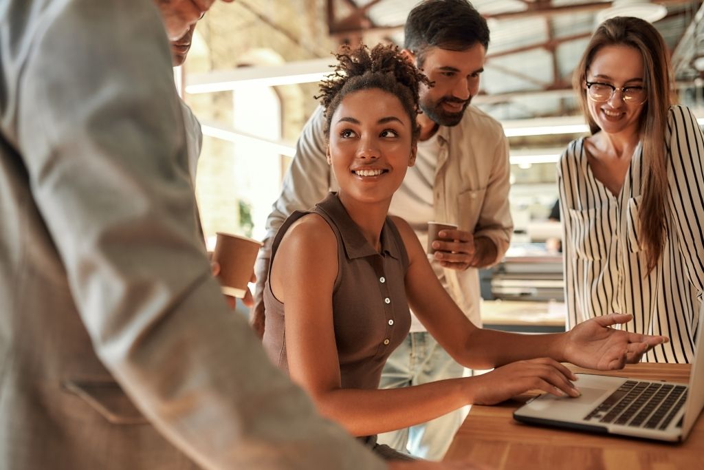 rupo de quatro profissionais diversos, incluindo homens e mulheres, em um ambiente de escritório, sorrindo e conversando enquanto analisam os desafios do RH. e possíveis soluções um laptop, representando um ambiente de trabalho colaborativo e comunicação dentro de um ambiente corporativo.