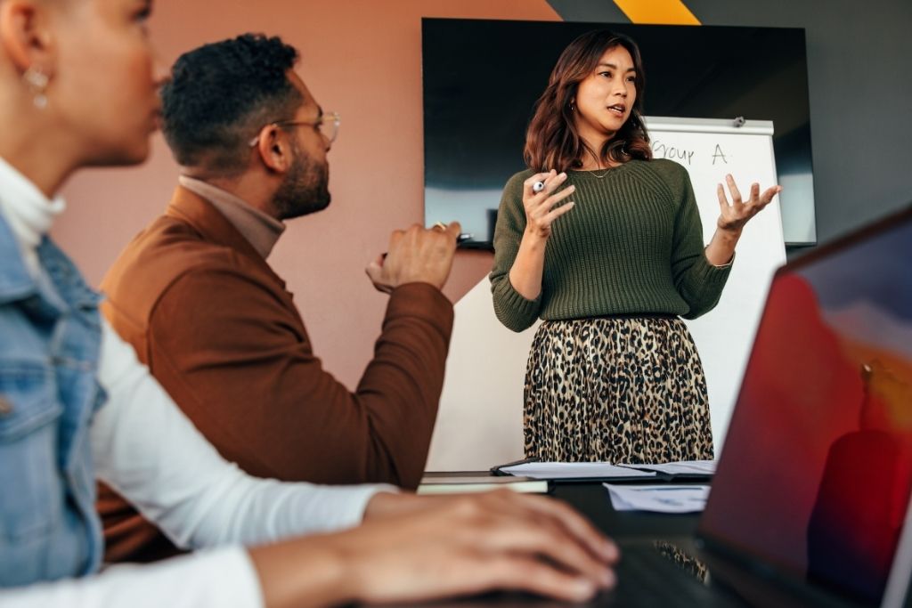 Uma mulher com cabelos castanhos e blusa verde gesticula durante uma reunião estratégica enquanto apresenta dados sobre KPIs de RH em um quadro branco. Ela está de pé diante de dois colegas que aparecem em primeiro plano, um homem de óculos e uma mulher de colete jeans, em um ambiente de escritório moderno e iluminado que sugere uma tomada de decisão baseada em indicadores.