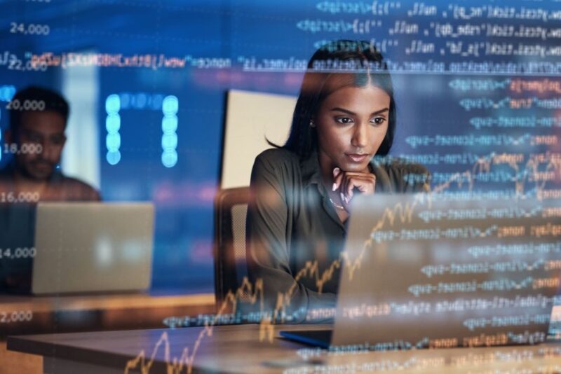 Uma mulher negra jovem com expressão concentrada trabalha em seu notebook em um escritório moderno. Sobreposta à cena, há uma camada digital transparente que exibe diversos gráficos de linha e blocos de código de programação, simbolizando a aplicação prática de people analytics no monitoramento de dados em tempo real. Ao fundo, um colega de trabalho aparece levemente desfocado, reforçando o ambiente corporativo tecnológico.