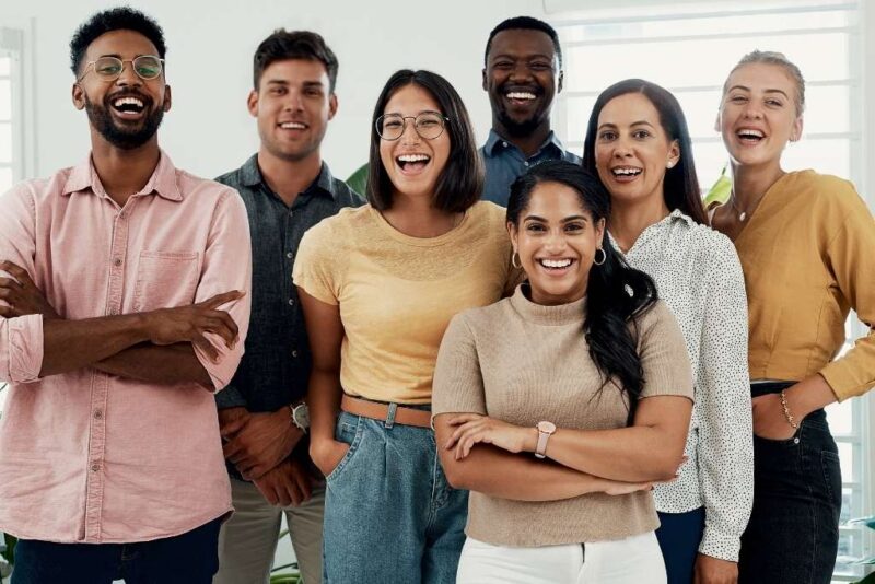Uma fotografia vibrante de um grupo diversificado de sete colegas de trabalho, homens e mulheres de diferentes etnias, sorrindo e rindo juntos em um ambiente de escritório iluminado. Eles vestem roupas casuais e demonstram uma postura relaxada e amigável, exemplificando o bem-estar no trabalho através da integração e da felicidade genuína da equipe no ambiente corporativo.