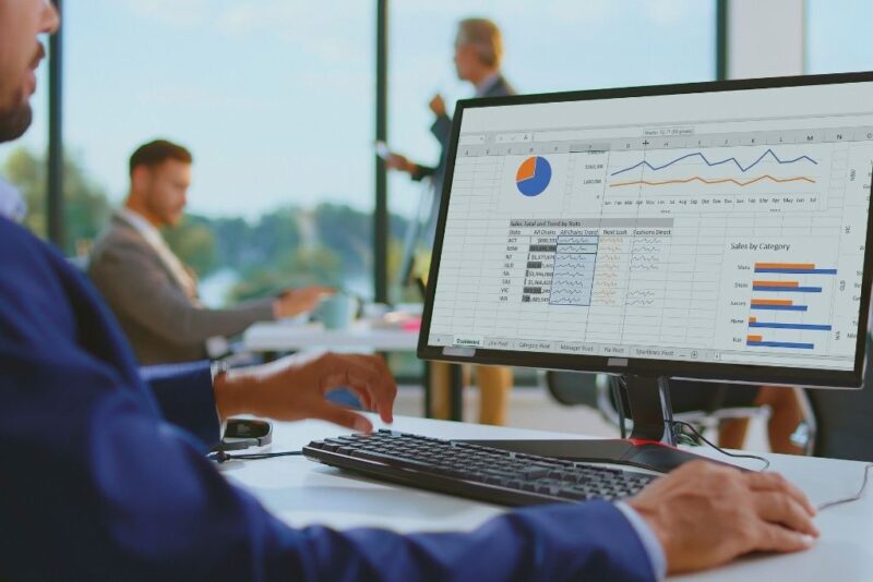 Homem sentado em um escritório moderno analisando planilhas de despesas operacionais em um monitor de computador. A tela exibe diversos gráficos de pizza, gráficos de linha e tabelas detalhadas com indicadores financeiros organizados por categorias e estados. Ao fundo, o ambiente corporativo mostra outros profissionais trabalhando, sugerindo um processo de gestão estratégica e monitoramento de custos empresariais.