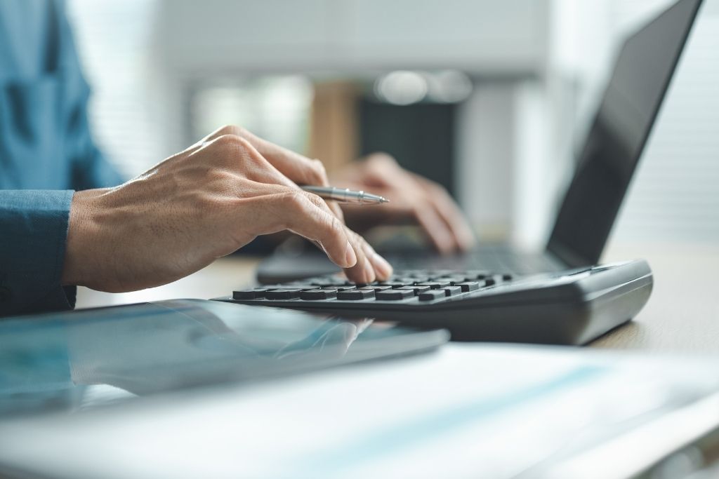 Close das mãos de um profissional utilizando uma calculadora financeira e um notebook para realizar o levantamento de despesas operacionais de uma empresa. A imagem foca na ação de calcular custos e gerir o fluxo de caixa, com documentos e um tablet dispostos sobre a mesa de trabalho em um ambiente de escritório iluminado. A composição enfatiza o rigor técnico necessário para a contabilidade e o planejamento financeiro organizacional.