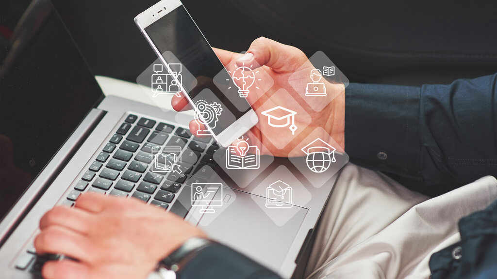 Pessoa utilizando celular e notebook para estudar educação corporativa de forma mobile, enquanto diversos ícones digitais ilustram a imagem.
