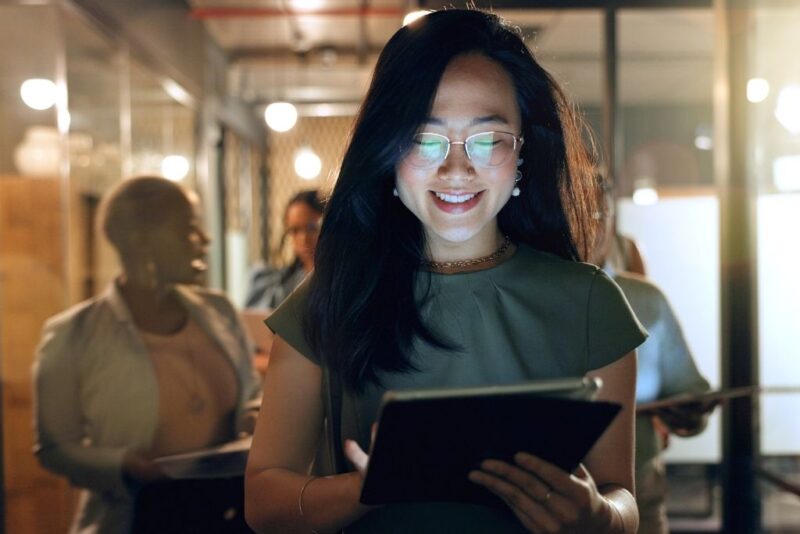 Uma mulher de óculos e cabelos escuros sorri enquanto utiliza um tablet em um ambiente de escritório moderno e bem iluminado. Ao fundo, outras colegas de trabalho aparecem levemente desfocadas, sugerindo um ambiente corporativo dinâmico e colaborativo. A imagem ilustra o uso da tecnologia e da mobilidade em processos de gestão de pessoas e demonstra na prática como medir a satisfação dos colaboradores através de ferramentas digitais e pesquisas de clima organizacional em tempo real.
