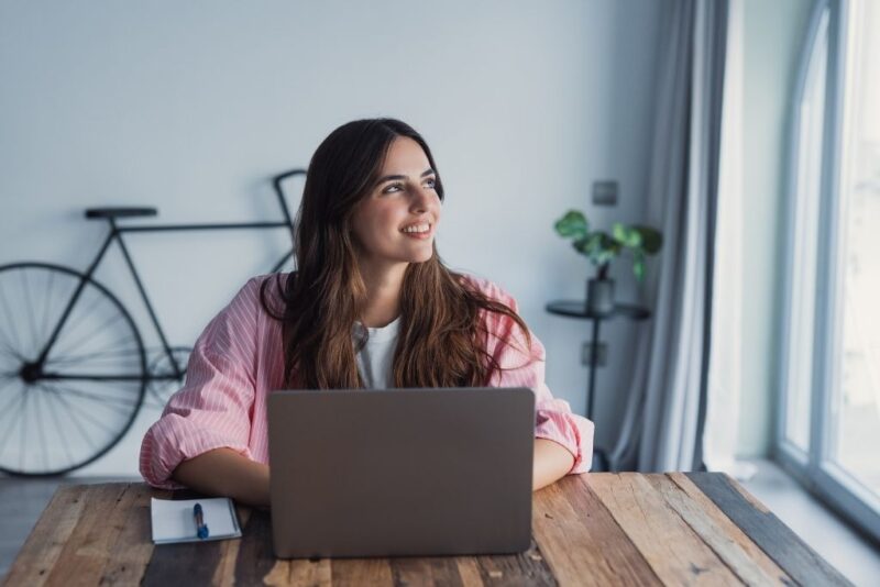 Uma mulher jovem de cabelos longos e castanhos sorri enquanto olha para o lado com um ar pensativo e otimista. Ela está sentada à frente de um laptop em uma mesa de madeira rústica, sugerindo um ambiente de trabalho confortável. Ao lado do computador, há um pequeno bloco de notas e uma caneta. No fundo do ambiente, é possível ver parte de uma bicicleta e uma planta, reforçando a ideia de um profissional que recebe ajuda de custo para manter sua estrutura de trabalho remoto com qualidade.