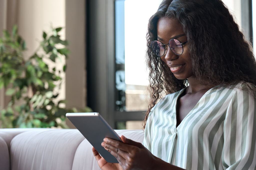 Uma mulher negra com cabelos longos e ondulados usa óculos e sorri suavemente enquanto lê um livro com um e-reader em um ambiente iluminado. Ela demonstra foco e satisfação, exemplificando um dos usos possíveis do Vale Cultura, para comprar livros digitais.
