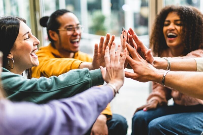Fotografia em close de um grupo diverso de quatro jovens adultos sorrindo e celebrando a cultura de diversidade e inclusão no ambiente de trabalho. A imagem foca nas mãos do grupo unidas ao centro em um gesto de "high five" coletivo, simbolizando colaboração e pertencimento. Entre os participantes, vê-se uma mulher branca à esquerda, um homem de traços asiáticos ao fundo e uma mulher negra à direita, todos expressando alegria e entusiasmo em um espaço bem iluminado.