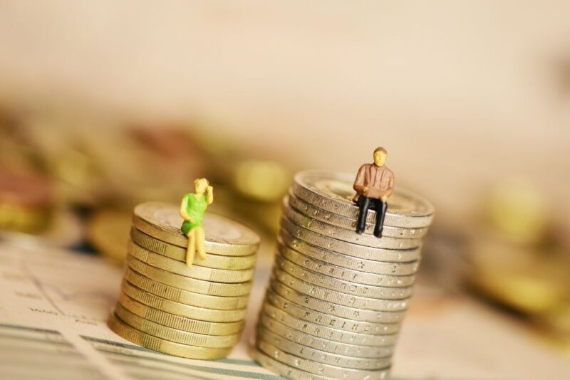 Uma fotografia conceitual com profundidade de campo reduzida que ilustra a falta de equidade salarial entre gêneros. Em primeiro plano sobre uma superfície com gráficos de desempenho, há duas pilhas de moedas de tamanhos diferentes. À esquerda, uma pilha menor com uma miniatura de mulher sentada no topo em posição reflexiva. À direita, uma pilha significativamente mais alta com uma miniatura de homem sentado de forma relaxada. O cenário ao fundo está desfocado, enfatizando o contraste visual entre o acúmulo de riqueza e a disparidade de rendimentos representada pelas alturas desiguais das moedas.