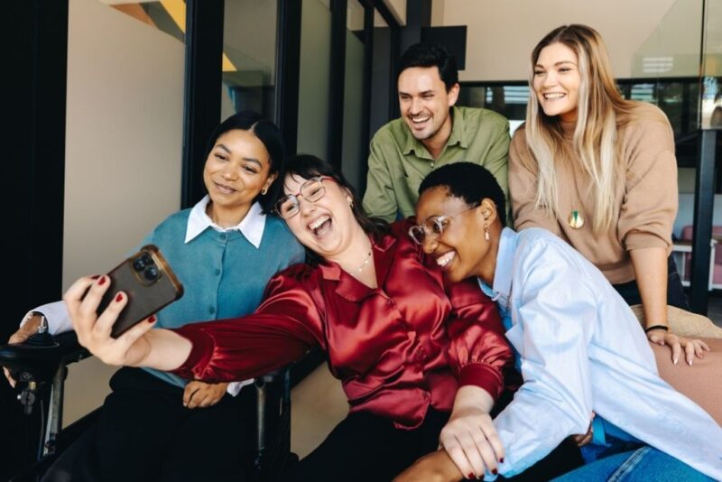 Uma equipe diversificada de cinco colegas de trabalho sorri e celebra junta enquanto uma mulher em primeiro plano segura um celular para tirar uma selfie do grupo. O clima é de extrema alegria e união, exemplificando o sucesso de campanhas e ideias de premiação para funcionários que buscam promover a integração e o reconhecimento em um ambiente corporativo moderno e inclusivo.