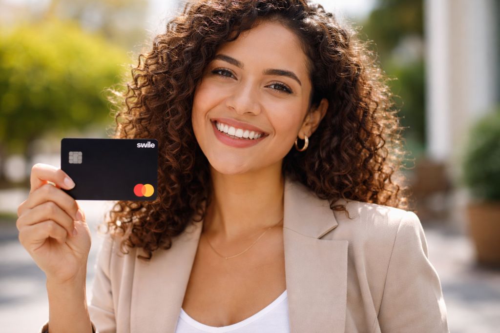 Uma jovem mulher brasileira com cabelos cacheados sorri com confiança para a câmera enquanto segura um cartão de benefícios preto da Swile, com a bandeira Mastercard. A foto em plano médio destaca o reconhecimento profissional em um ambiente externo iluminado, sugerindo a eficácia de utilizar benefícios flexíveis como uma das melhores ideias de premiação para funcionários para elevar a satisfação e a liberdade de escolha da equipe.