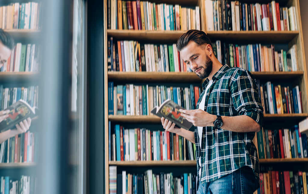 Um homem jovem com barba aparada e camisa xadrez está de pé em uma livraria ou biblioteca, folheando atentamente um livro. Ao fundo, diversas estantes repletas de obras coloridas reforçam o incentivo à leitura e o acesso ao conhecimento. Este cenário representa o benefício de adquirir novas obras literárias por meio do incentivo oferecido pelo Vale Cultura.