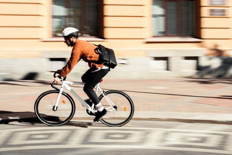Fotografia em ângulo lateral capturando um ciclista em movimento por uma rua urbana. O homem utiliza um capacete de proteção branco, blusa de lã alaranjada e carrega uma mochila transversal preta nas costas. A imagem utiliza um efeito de desfoque de movimento no plano de fundo, que apresenta edifícios de arquitetura clássica. O registro enfatiza a agilidade e a eficiência do uso da bicicleta como uma escolha prática de mobilidade sustentável nos grandes centros.