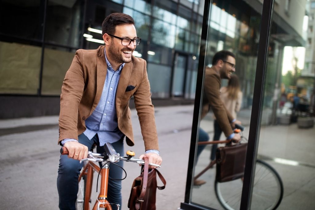 Um homem jovem executivo de barba e óculos sorri alegremente enquanto pedala uma bicicleta urbana em uma rua da cidade. Ele veste um paletó marrom sobre uma camisa social azul, transmitindo uma imagem de profissionalismo e bem-estar. O reflexo do ciclista é visível na no vidro de um ponto de ônibus moderno ao lado. A cena ilustra como a mobilidade sustentável pode ser integrada à rotina de trabalho, promovendo disposição e produtividade no trajeto corporativo.