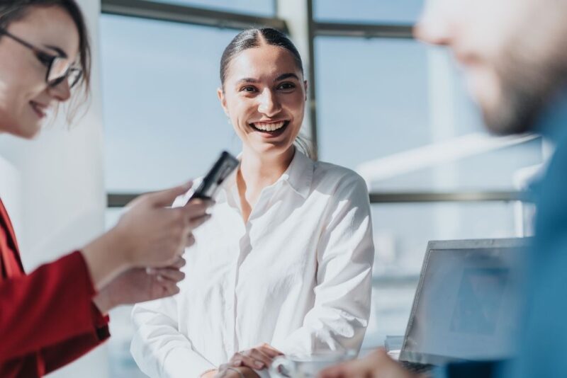 Uma fotografia em close de três colegas de trabalho em um ambiente de escritório moderno e iluminado pelo sol. No centro, uma mulher sorri abertamente enquanto participa de uma conversa descontraída, transmitindo a satisfação de uma colaboradora que conta com um pacote de benefícios atrativo. À esquerda, outra profissional segura um smartphone, e à direita, parte de um colega e seu notebook aparecem em primeiro plano, reforçando o clima de colaboração e bem-estar no ambiente corporativo.