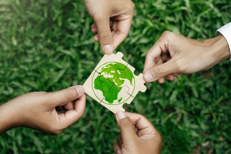A imagem apresenta um plano detalhado de quatro mãos que seguram e unem peças de um quebra-cabeça de madeira sobre um fundo de grama verde. As peças montadas formam o desenho do globo terrestre na cor verde, simbolizando a união e o esforço coletivo necessários para implementar práticas ESG com foco na preservação ambiental e na sustentabilidade global.
