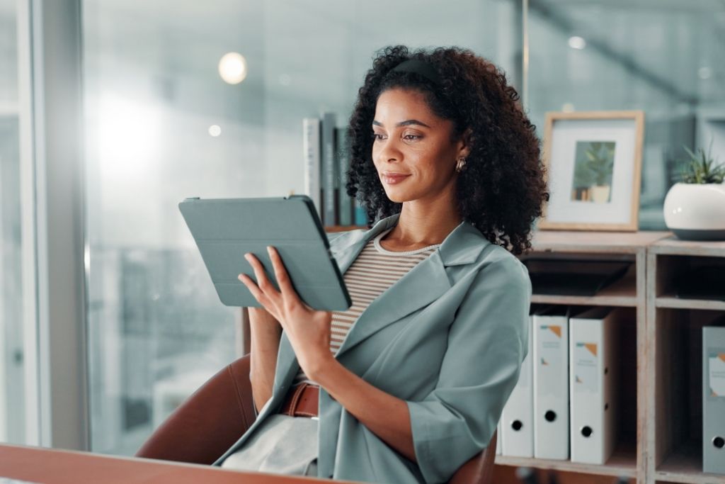 Uma mulher jovem com cabelos cacheados e sorridente veste um blazer azul claro e manuseia um tablet em um ambiente de escritório moderno e bem iluminado. Ela demonstra foco e tranquilidade ao analisar dados digitais, representando a etapa de monitoramento e a governança corporativa essenciais para o sucesso das práticas ESG dentro de uma organização.