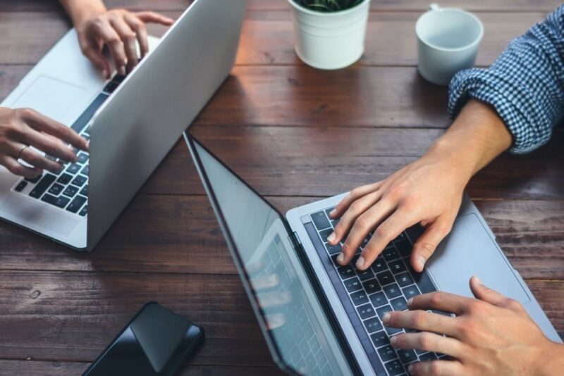 Vista superior em close de duas pessoas trabalhando em notebooks sobre uma mesa de madeira rústica, representando o ambiente corporativo onde se discute remuneração e salário de colaboradores. É possível ver apenas as mãos dos profissionais digitando nos teclados prateados, além de um smartphone e uma caneca branca ao lado, simbolizando a rotina produtiva e o desempenho profissional.