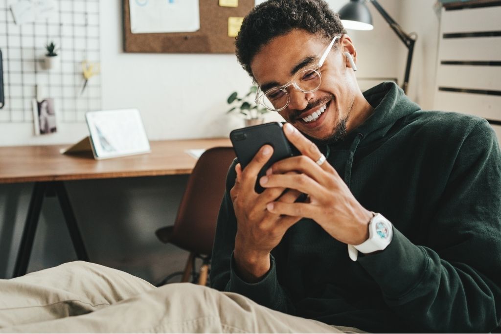 Um homem jovem de pele negra com óculos e fones de ouvido sorri alegremente enquanto olha para a tela de seu celular em um escritório moderno, celebrando possivelmente boas notícias sobre sua remuneração e salário. Ao fundo, o ambiente de trabalho organizado conta com uma mesa de madeira e um mural de recados, transmitindo uma sensação de satisfação e bem-estar no emprego.