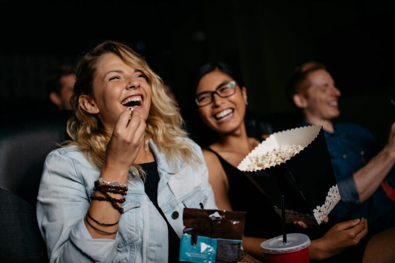 Uma mulher loira sorri com entusiasmo e gargalha enquanto come pipoca em uma sala de cinema escura. Ao lado dela, outros jovens também se divertem e dão risada, criando um ambiente descontraído de lazer. A cena ilustra um momento clássico de entretenimento que pode ser aproveitado com o uso do Vale Cultura para acesso a entretenimento cultural.