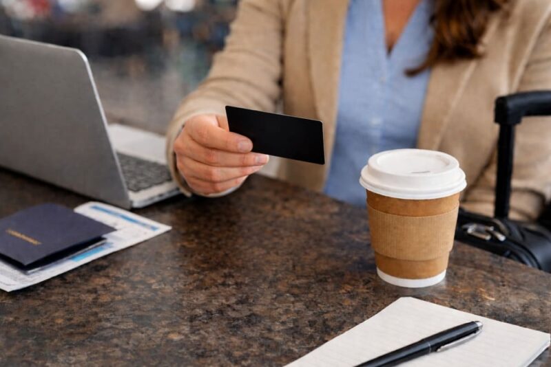 Profissional segurando cartão corporativo preto, enquanto está sentada com mala ao lado, notebook, caderno, caneta, passagem e café na mesa a frente.