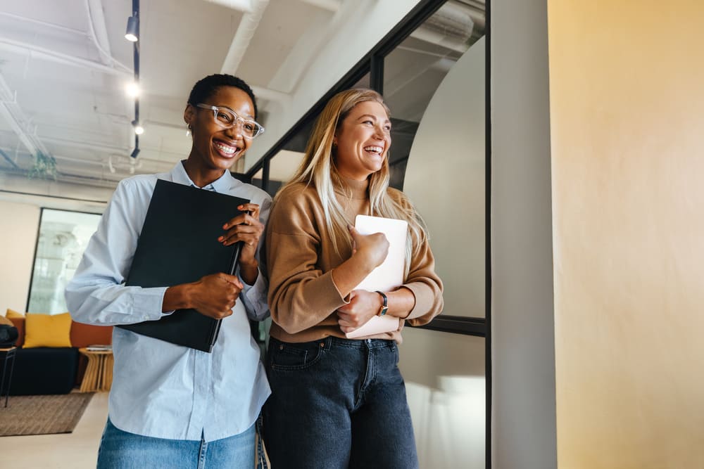Duas colaboradoras sorrindo em escritório corporativo, representando como iniciativas de bem-estar, como o vale-academia, contribuem para uma experiência mais positiva e engajadora no ambiente de trabalho.