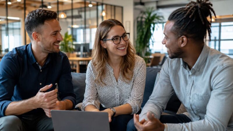 Três colegas de trabalho sentados próximos e sorrindo em ambiente corporativo, trocando percepções e reforçando a cultura do feedback na empresa.